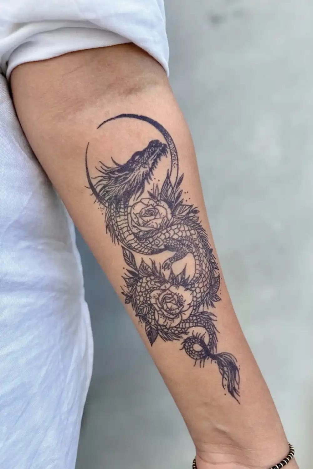 Forearm wearing a black temporary tattoo of a dragon wrapped around roses inside a crescent arc, white shirt