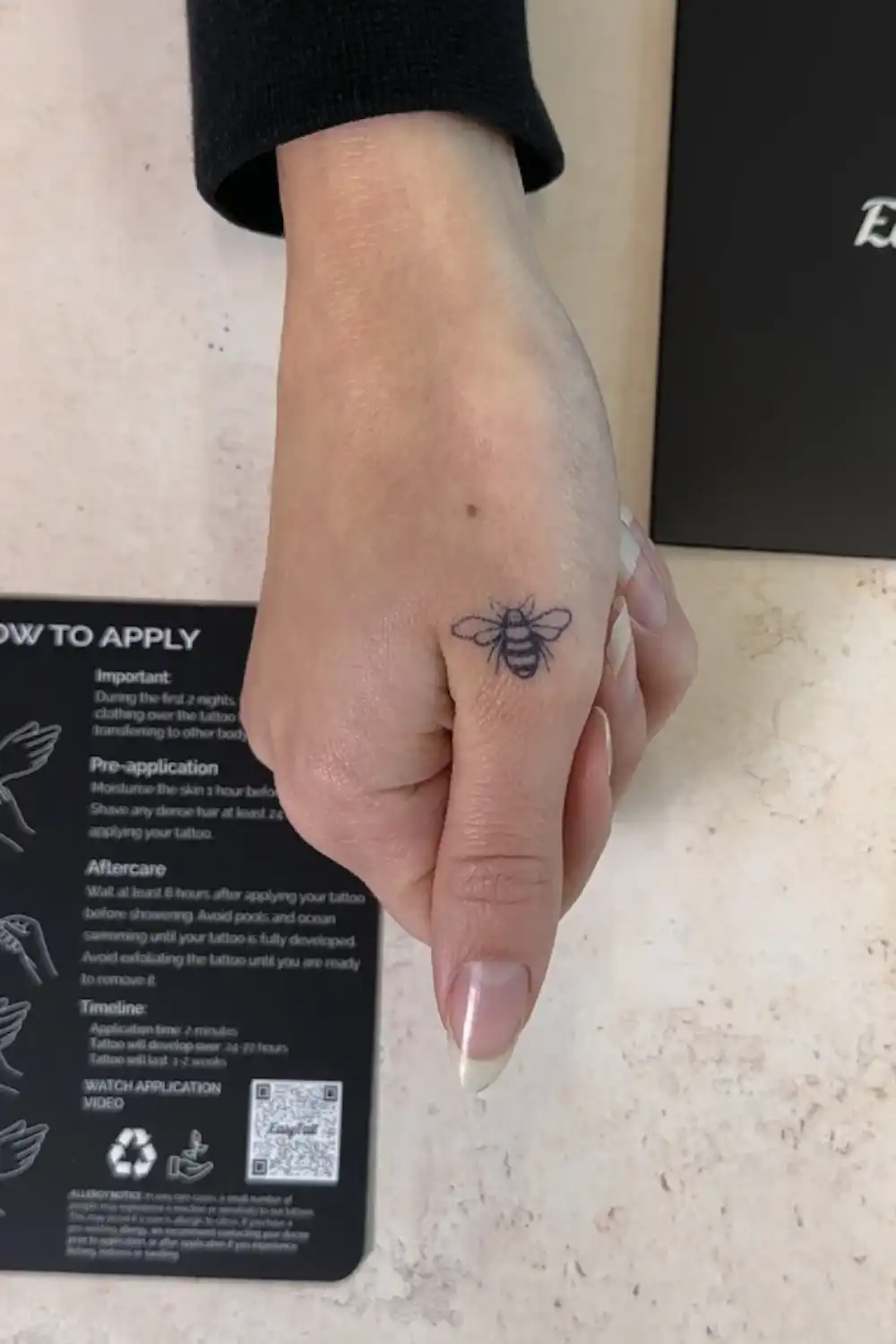 Small bee EasyTatt temporary tattoo applied to the base of a thumb, positioned next to the brand's black instruction card